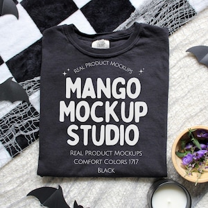 Op de afbeelding: Een zwart Comfort Colors 1717 t-shirt met de tekst "MANGO MOCKUP STUDIO" in het wit. Het shirt wordt gepresenteerd op een zwart-wit geruit oppervlak met decoratieve vleermuizen en een kleine houten kom met paarse bloemen.