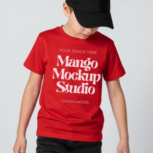 May include: A red t-shirt with white text that reads "YOUR DESIGN HERE Mango Mockup Studio Gildan 64000B". The shirt is short-sleeved and is being worn by a person wearing a black baseball cap.