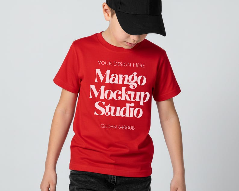 May include: A red t-shirt with white text that reads "YOUR DESIGN HERE Mango Mockup Studio Gildan 64000B". The shirt is short-sleeved and is being worn by a person wearing a black baseball cap.