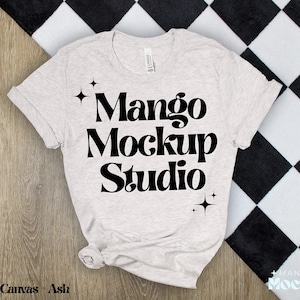 May include: A light grey t-shirt with black text that reads "Mango Mockup Studio" and two small stars on either side of the text. The t-shirt is laid flat on a black and white chequered surface.