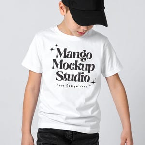 May include: A white t-shirt with the text "Mango Mockup Studio" in a bold, black font. The shirt is being worn by a person wearing a black baseball cap and dark jeans. The text "Your Design Here" is below the main text.