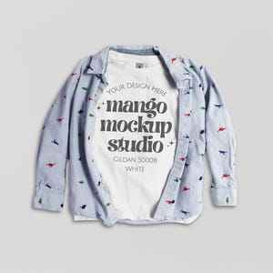 May include: A white Gildan 5000B t-shirt with the text "mango mockup studio" is layered under a light blue button-down shirt. The button-down shirt has a white stripe pattern and small dinosaur designs in various colours.