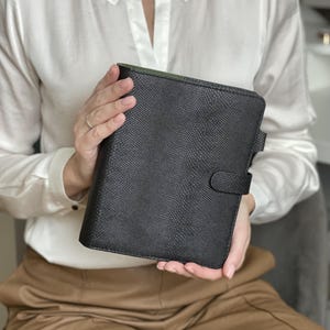 Pode incluir: Um planejador de couro preto texturizado com fecho de press&atilde;o. O planejador &eacute; segurado por uma pessoa vestindo uma blusa branca e cal&ccedil;as bege. O interior do planejador &eacute; verde.