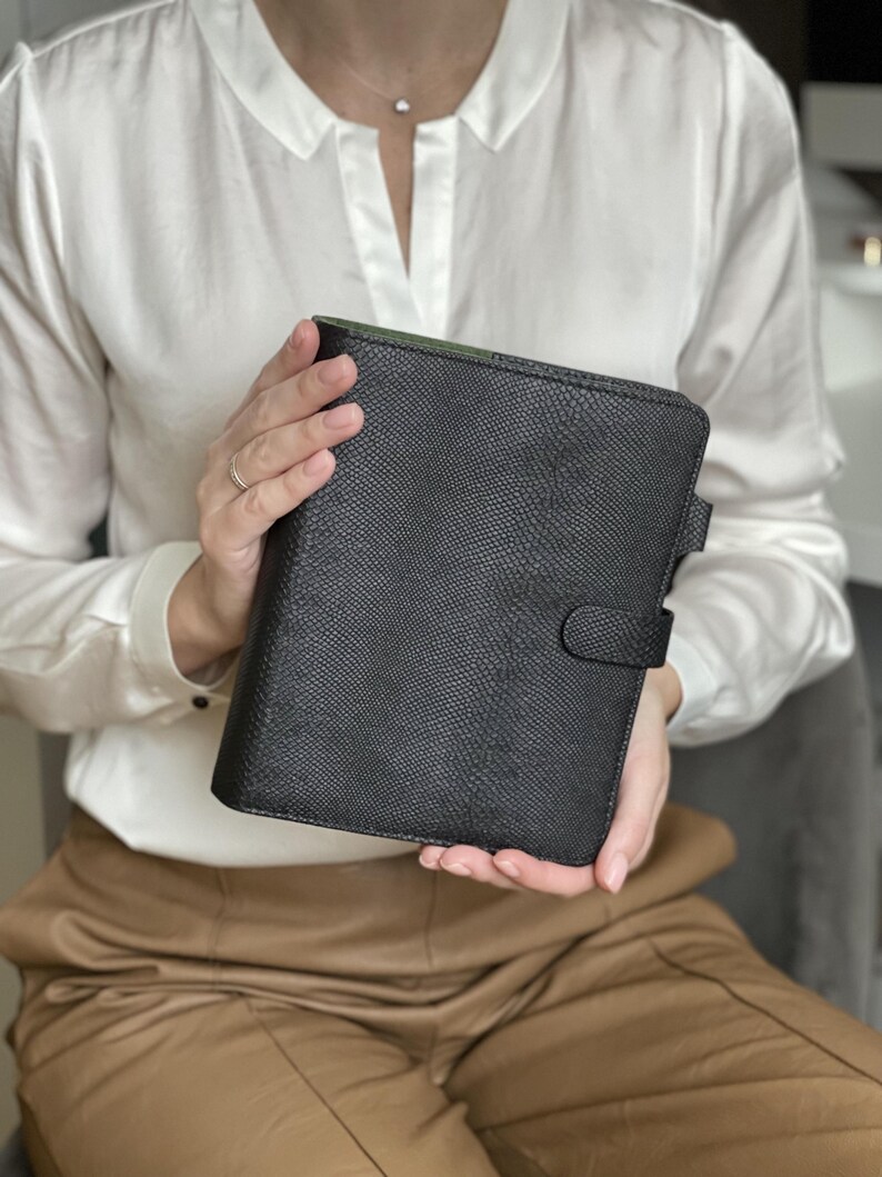 Pode incluir: Um planejador de couro preto texturizado com fecho de press&atilde;o. O planejador &eacute; segurado por uma pessoa vestindo uma blusa branca e cal&ccedil;as bege. O interior do planejador &eacute; verde.