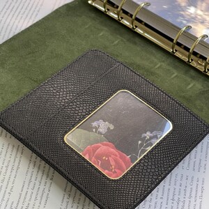 Pode incluir: Carteira de couro preta com um padr&atilde;o texturizado de pele de cobra. A carteira apresenta uma janela transparente com um desenho floral de uma rosa vermelha e pequenas flores roxas. A carteira est&aacute; sobre uma superf&iacute;cie verde.