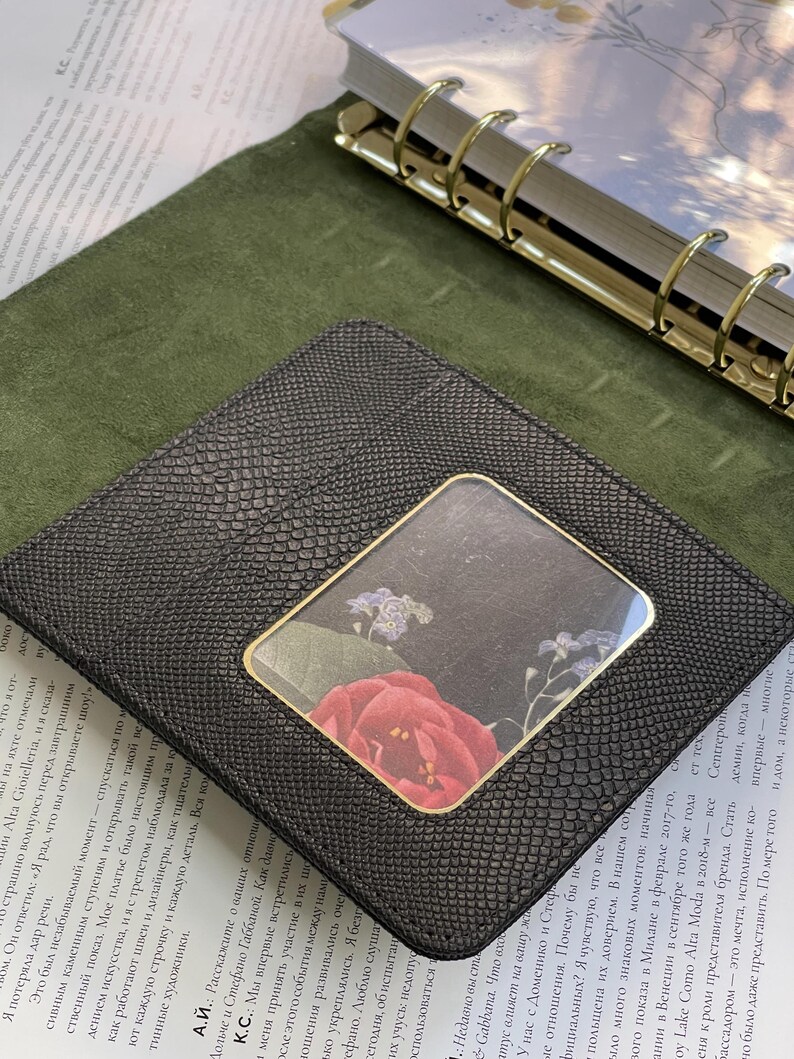 Pode incluir: Carteira de couro preta com um padr&atilde;o texturizado de pele de cobra. A carteira apresenta uma janela transparente com um desenho floral de uma rosa vermelha e pequenas flores roxas. A carteira est&aacute; sobre uma superf&iacute;cie verde.