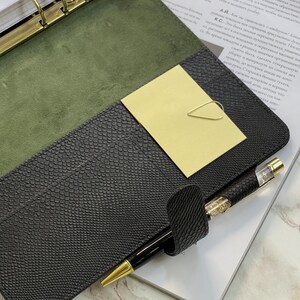 Pode incluir: Um planejador de couro preto com detalhes dourados, aberto para revelar um interior verde e uma caneta dourada. Um clip de papel dourado segura um cart&atilde;o amarelo. O planejador est&aacute; em cima de uma revista.