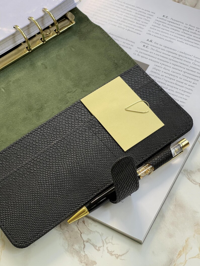 Pode incluir: Um planejador de couro preto com detalhes dourados, aberto para revelar um interior verde e uma caneta dourada. Um clip de papel dourado segura um cart&atilde;o amarelo. O planejador est&aacute; em cima de uma revista.