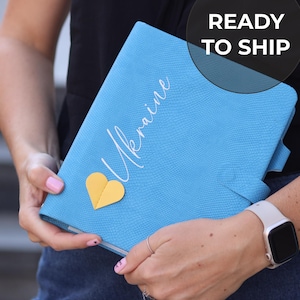Peut inclure: Un cahier bleu texturé avec le mot "Ukraine" en écriture cursive et un cœur jaune. Le cahier est ouvert, avec le texte "READY TO SHIP" dans un cercle noir en haut à droite.