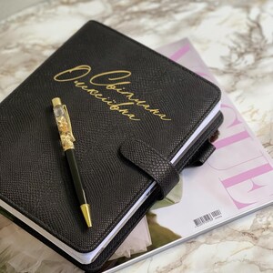 Pode incluir: Di&aacute;rio de couro preto com letras douradas e caneta a condizer. O di&aacute;rio tem uma capa texturizada e um fecho de fivela. A caneta tem uma ponta dourada e &eacute; preenchida com flocos de ouro. O di&aacute;rio est&aacute; sobre uma revista.