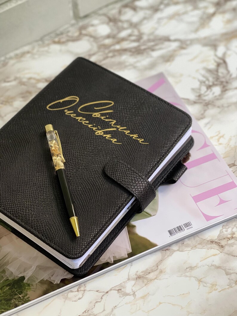 Pode incluir: Di&aacute;rio de couro preto com letras douradas e caneta a condizer. O di&aacute;rio tem uma capa texturizada e um fecho de fivela. A caneta tem uma ponta dourada e &eacute; preenchida com flocos de ouro. O di&aacute;rio est&aacute; sobre uma revista.