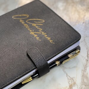 Pode incluir: Di&aacute;rio de couro preto com padr&atilde;o de pele de cobra e letras douradas. Uma caneta preta e dourada est&aacute; presa na lombada. O di&aacute;rio &eacute; fechado com uma tira de couro a condizer.