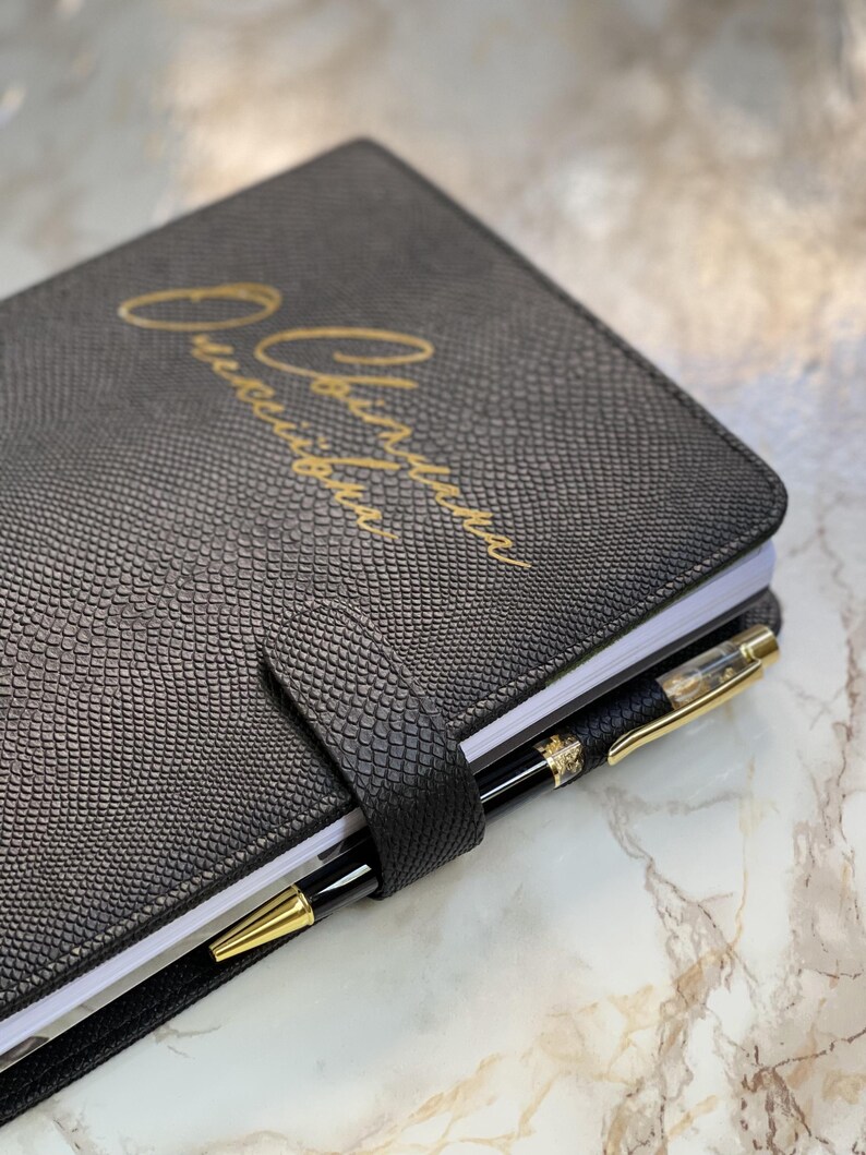 Pode incluir: Di&aacute;rio de couro preto com padr&atilde;o de pele de cobra e letras douradas. Uma caneta preta e dourada est&aacute; presa na lombada. O di&aacute;rio &eacute; fechado com uma tira de couro a condizer.