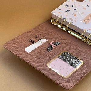 Peut inclure: Un agenda en similicuir marron avec des anneaux dorés et une couverture à motifs. L'agenda est ouvert, révélant des poches avec une carte blanche, une trombone rose et une fenêtre transparente avec un motif noir et blanc.