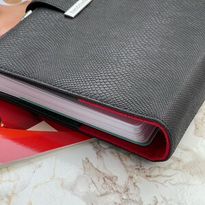 Pode incluir: Um planejador de couro preto texturizado com forro interno vermelho. O planejador &eacute; fechado com uma al&ccedil;a e um fecho de metal prateado que diz "handmade". O planejador est&aacute; sobre uma revista com fundo de m&aacute;rmore.