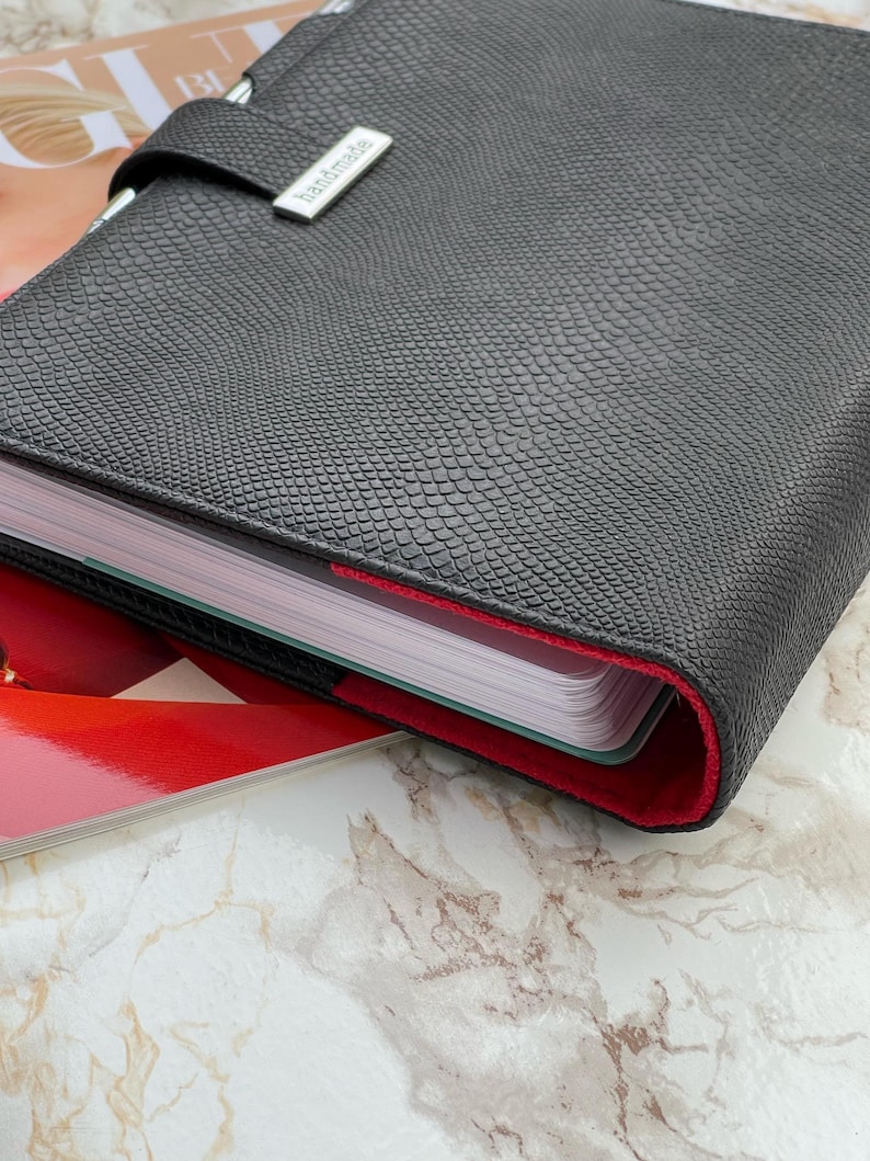 Pode incluir: Um planejador de couro preto texturizado com forro interno vermelho. O planejador &eacute; fechado com uma al&ccedil;a e um fecho de metal prateado que diz "handmade". O planejador est&aacute; sobre uma revista com fundo de m&aacute;rmore.