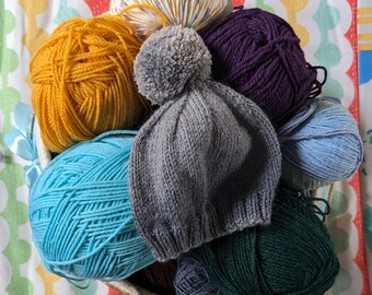 May include: A basket filled with colourful yarn balls and a grey knitted hat with a grey pom-pom. The yarn balls are in shades of blue, yellow, purple, and white.