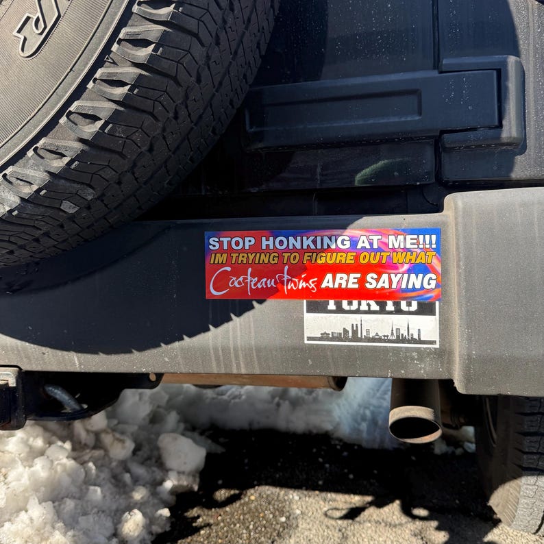 May include: A bumper sticker with the text "STOP HONKING AT ME!!! IM TRYING TO FIGURE OUT WHAT Cocteau Twins ARE SAYING" in white and red lettering. Below is a smaller sticker with a city skyline.Stop honking at me funny trendy band bumper sticker, music meme bumper sticker, funny trendy sticker decal, internet humor first car gift on laptop as laptop sticker decal