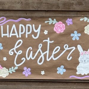 May include: A brown paper banner with the words "HAPPY Easter" in white script. The banner is decorated with pastel flowers, a bunny, and a purple bow. The banner is mounted on a wooden background.