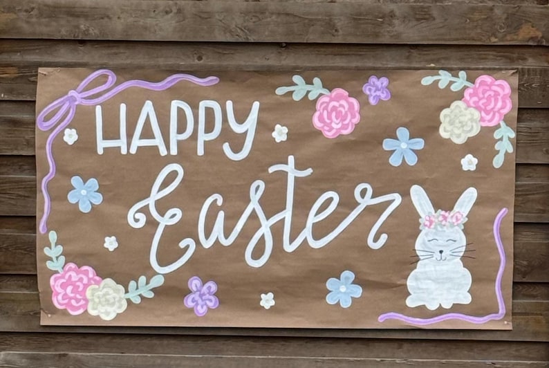 May include: A brown paper banner with the words "HAPPY Easter" in white script. The banner is decorated with pastel flowers, a bunny, and a purple bow. The banner is mounted on a wooden background.