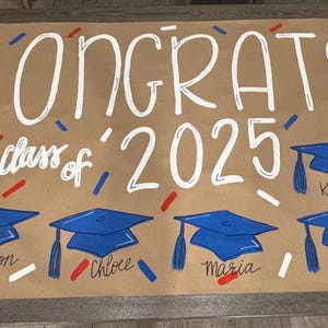 May include: A brown paper banner with the word "CONGRATS" in white script, along with "class of 2025". Blue graduation caps are painted below the text, each with a student's name. Red, white, and blue confetti is scattered around.