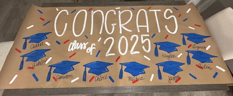 May include: A brown paper banner with the word "CONGRATS" in white script, along with "class of 2025". Blue graduation caps are painted below the text, each with a student's name. Red, white, and blue confetti is scattered around.
