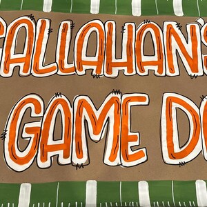 May include: A brown banner with the words "CALLAHAN'S 1ST GAME DAY" in orange and white lettering. A football and a Longhorn silhouette are also featured. The banner has a green and white striped border, suggesting a sports theme.