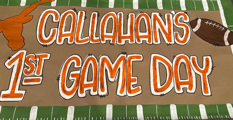 May include: A brown banner with the words "CALLAHAN'S 1ST GAME DAY" in orange and white lettering. A football and a Longhorn silhouette are also featured. The banner has a green and white striped border, suggesting a sports theme.