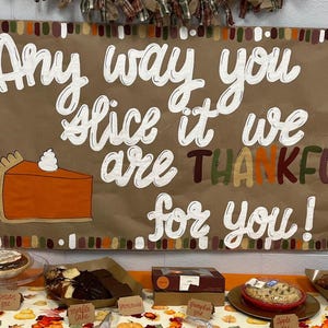 May include: A brown poster with the text "Any way you slice it we are THANKFUL for you!" and a slice of pumpkin pie. Various pies and cakes are displayed on a table below the poster.