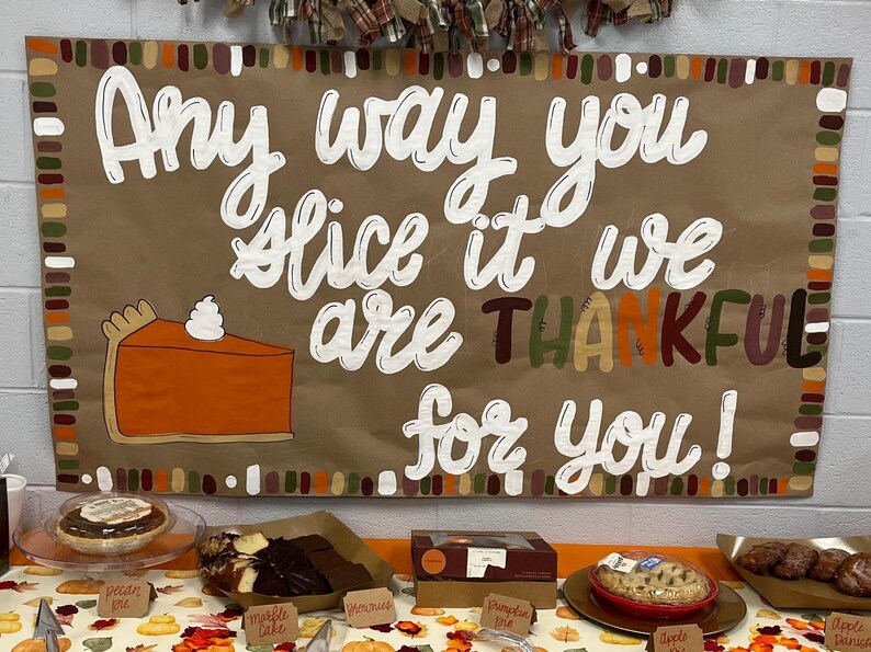 May include: A brown poster with the text "Any way you slice it we are THANKFUL for you!" and a slice of pumpkin pie. Various pies and cakes are displayed on a table below the poster.