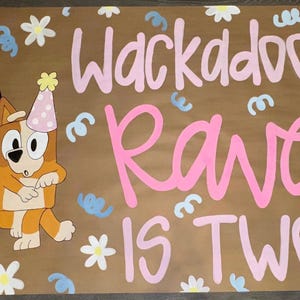 May include: A brown poster with the words "Wackadoo! Rawe is TWO!" in pink and white lettering. Two cartoon dogs wearing party hats are on the left. The poster is decorated with white flowers and blue confetti.