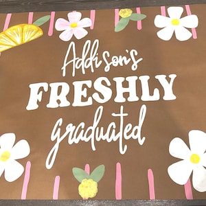May include: A brown graduation sign with the text "Addison's FRESHLY graduated" in white. The sign is decorated with white flowers, pink flowers, and yellow lemon slices. Pink vertical stripes are also present.