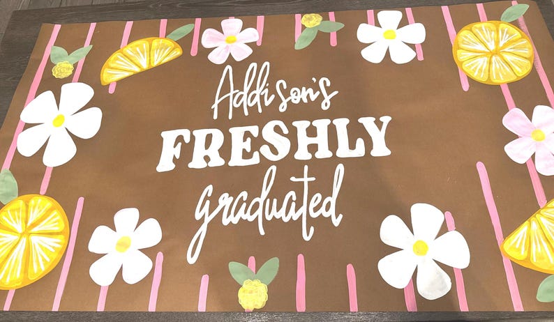 May include: A brown graduation sign with the text "Addison's FRESHLY graduated" in white. The sign is decorated with white flowers, pink flowers, and yellow lemon slices. Pink vertical stripes are also present.