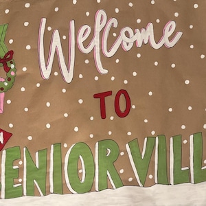 May include: A brown banner with the words "Welcome to Seniorville" in white and green. The banner features a Grinch character in a Santa hat, a signpost with arrows, and a wreath. White polka dots are scattered across the banner.