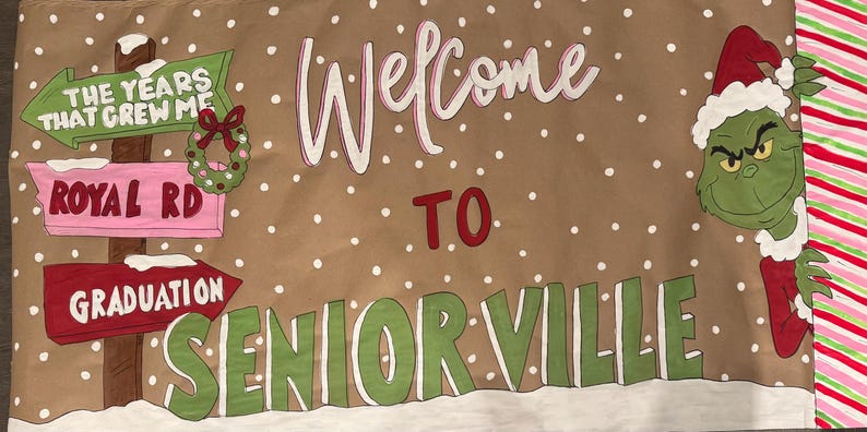 May include: A brown banner with the words "Welcome to Seniorville" in white and green. The banner features a Grinch character in a Santa hat, a signpost with arrows, and a wreath. White polka dots are scattered across the banner.