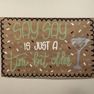 May include: A brown paper sign with the words "Say Say is just a tini bit older" in green and white lettering. A martini glass with olives is drawn on the right. The sign has a black and white scalloped border and confetti.