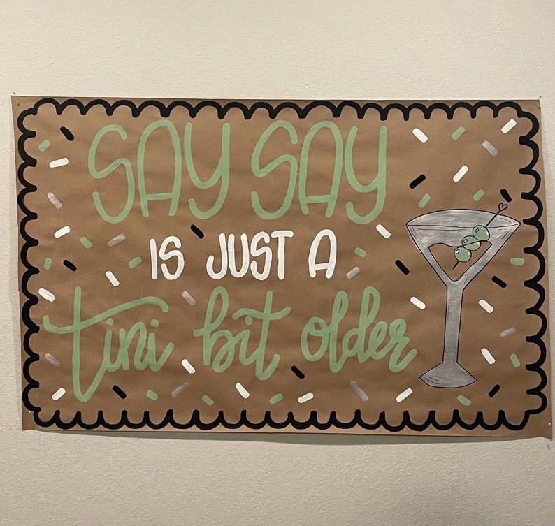 May include: A brown paper sign with the words "Say Say is just a tini bit older" in green and white lettering. A martini glass with olives is drawn on the right. The sign has a black and white scalloped border and confetti.