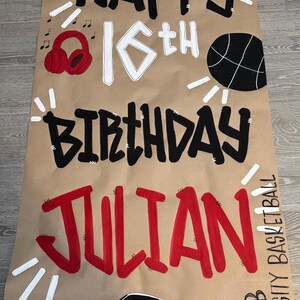 May include: A brown paper birthday banner with the words "HAPPY 16th BIRTHDAY JULIAN" in black and red lettering. The banner includes a basketball, headphones, a car, and the text "SEB VARSITY BASKETBALL".