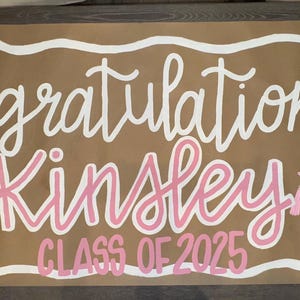 May include: A brown graduation banner with white and pink lettering. The banner says "Congratulations Kinsley CLASS OF 2025". It features pink bows, flowers, a diploma, and a graduation cap.