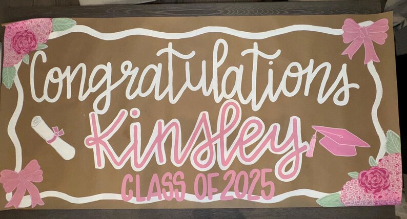 May include: A brown graduation banner with white and pink lettering. The banner says "Congratulations Kinsley CLASS OF 2025". It features pink bows, flowers, a diploma, and a graduation cap.
