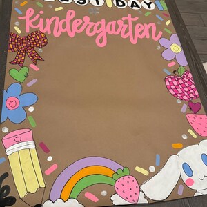 May include: A brown poster with the words "FIRST DAY Kindergarten" in pink and white. The poster is decorated with colorful illustrations of a bow, flowers, a pencil, a rainbow, an apple, a strawberry, and a cartoon bunny.
