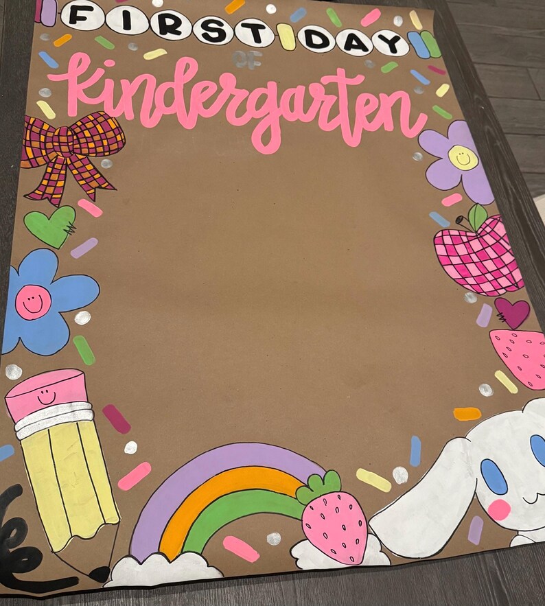May include: A brown poster with the words "FIRST DAY Kindergarten" in pink and white. The poster is decorated with colorful illustrations of a bow, flowers, a pencil, a rainbow, an apple, a strawberry, and a cartoon bunny.