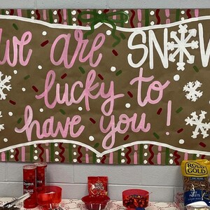May include: A brown sign with the words "we are lucky to have you!" in pink and white lettering, with snowflake accents. The sign is decorated with a white border and a green bow. Snacks are on a table below.