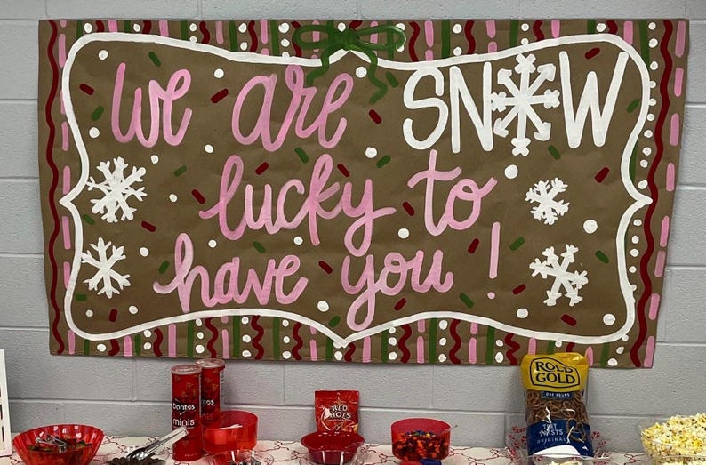 May include: A brown sign with the words "we are lucky to have you!" in pink and white lettering, with snowflake accents. The sign is decorated with a white border and a green bow. Snacks are on a table below.