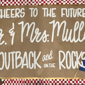 May include: A brown sign with white lettering and illustrations, including champagne glasses, wedding rings, and an Australian flag. The text reads "Cheers to the Future Mr. & Mrs. Mulloy Outback and on the Rocks."
