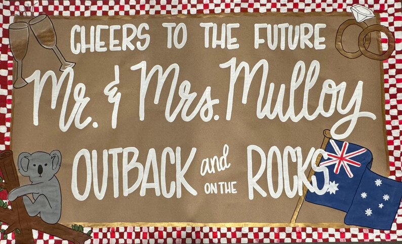 May include: A brown sign with white lettering and illustrations, including champagne glasses, wedding rings, and an Australian flag. The text reads "Cheers to the Future Mr. & Mrs. Mulloy Outback and on the Rocks."