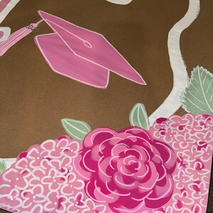 May include: A brown graduation-themed artwork featuring a pink mortarboard, tassel, and floral accents. The design includes a large pink rose and clusters of pink flowers, with white script lettering at the top. Green leaves add a touch of contrast.