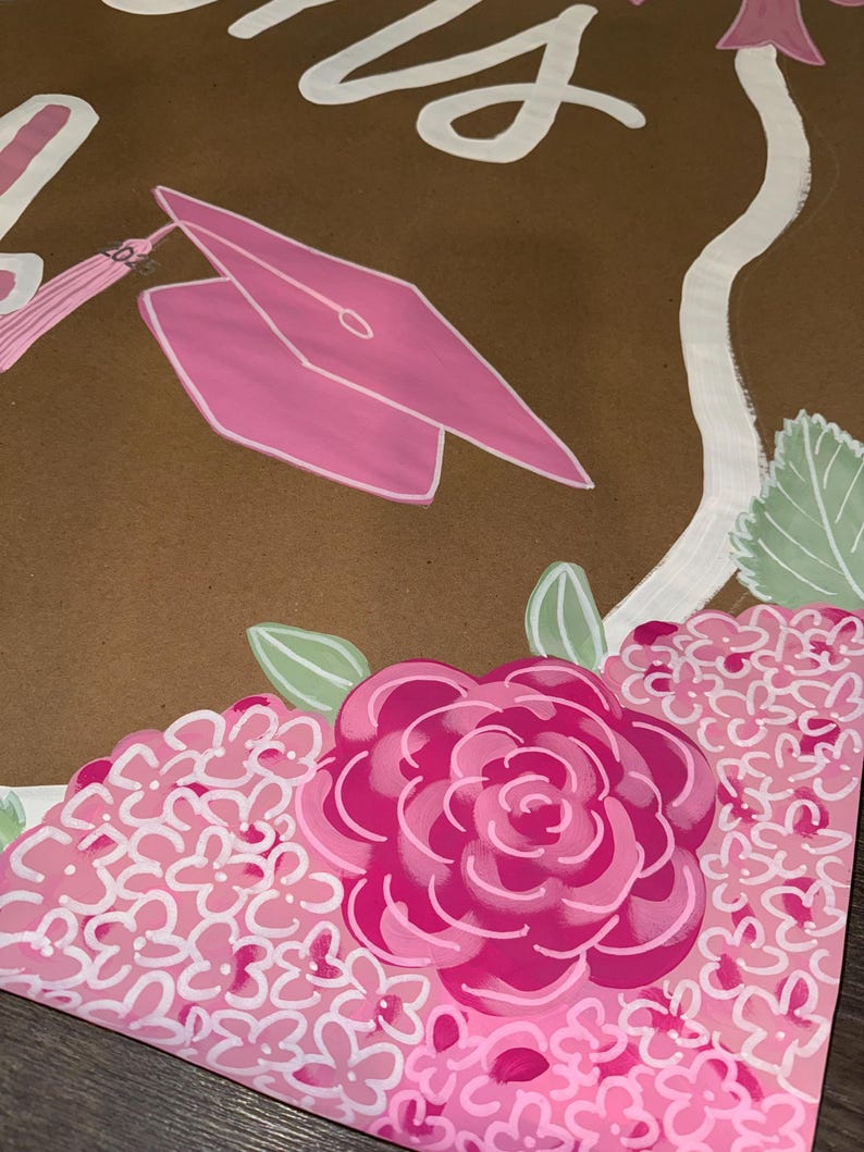 May include: A brown graduation-themed artwork featuring a pink mortarboard, tassel, and floral accents. The design includes a large pink rose and clusters of pink flowers, with white script lettering at the top. Green leaves add a touch of contrast.