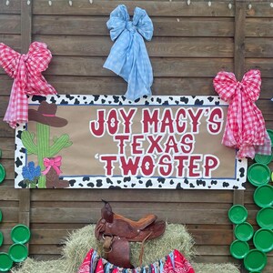 May include: A rustic wooden backdrop with a sign that reads "JOY MACY'S TEXAS TWOSTEP." The sign features cowboy-themed illustrations and is decorated with pink and blue bows. A saddle rests on a hay bale.