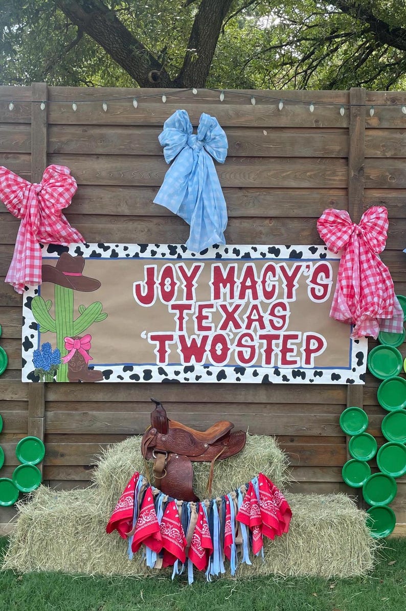 May include: A rustic wooden backdrop with a sign that reads "JOY MACY'S TEXAS TWOSTEP." The sign features cowboy-themed illustrations and is decorated with pink and blue bows. A saddle rests on a hay bale.